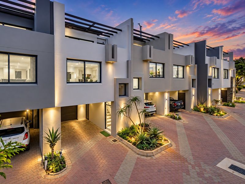 A modern multi-level townhouse complex featuring a paved driveway, integrated garages, and contemporary architectural facade with integrated exterior lighting.