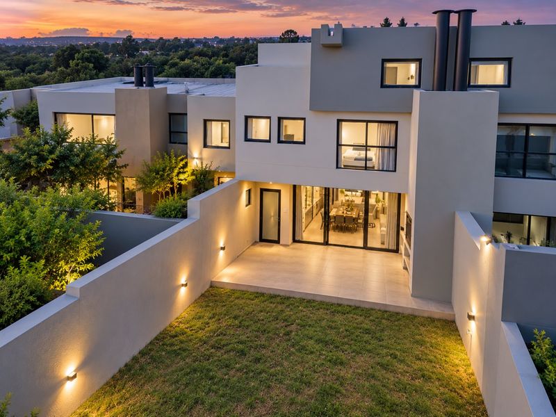 Modern rear elevation featuring a tiled patio, built-in fireplace, and landscaped garden, illuminated by integrated wall lighting at dusk.