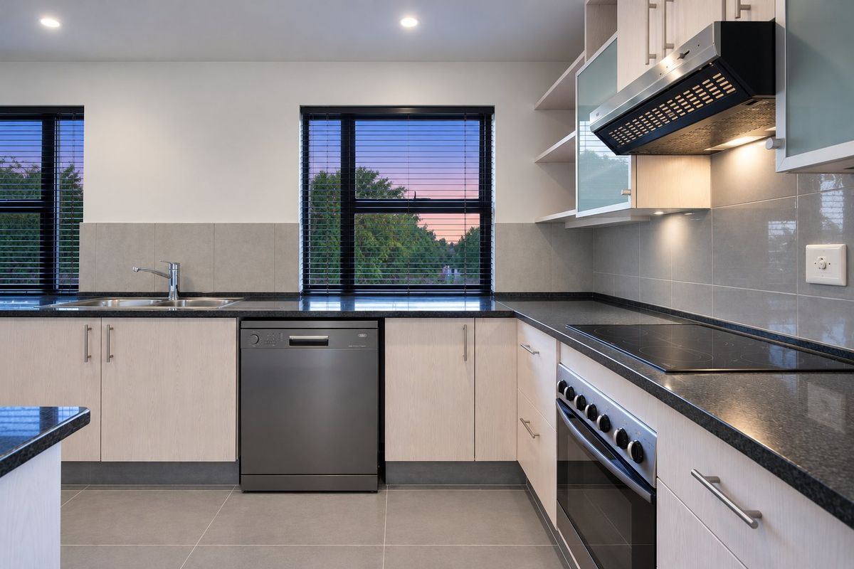 Modern kitchen featuring light wood cabinetry, dark granite countertops, integrated appliances, and a tiled backsplash with views of the outdoors.