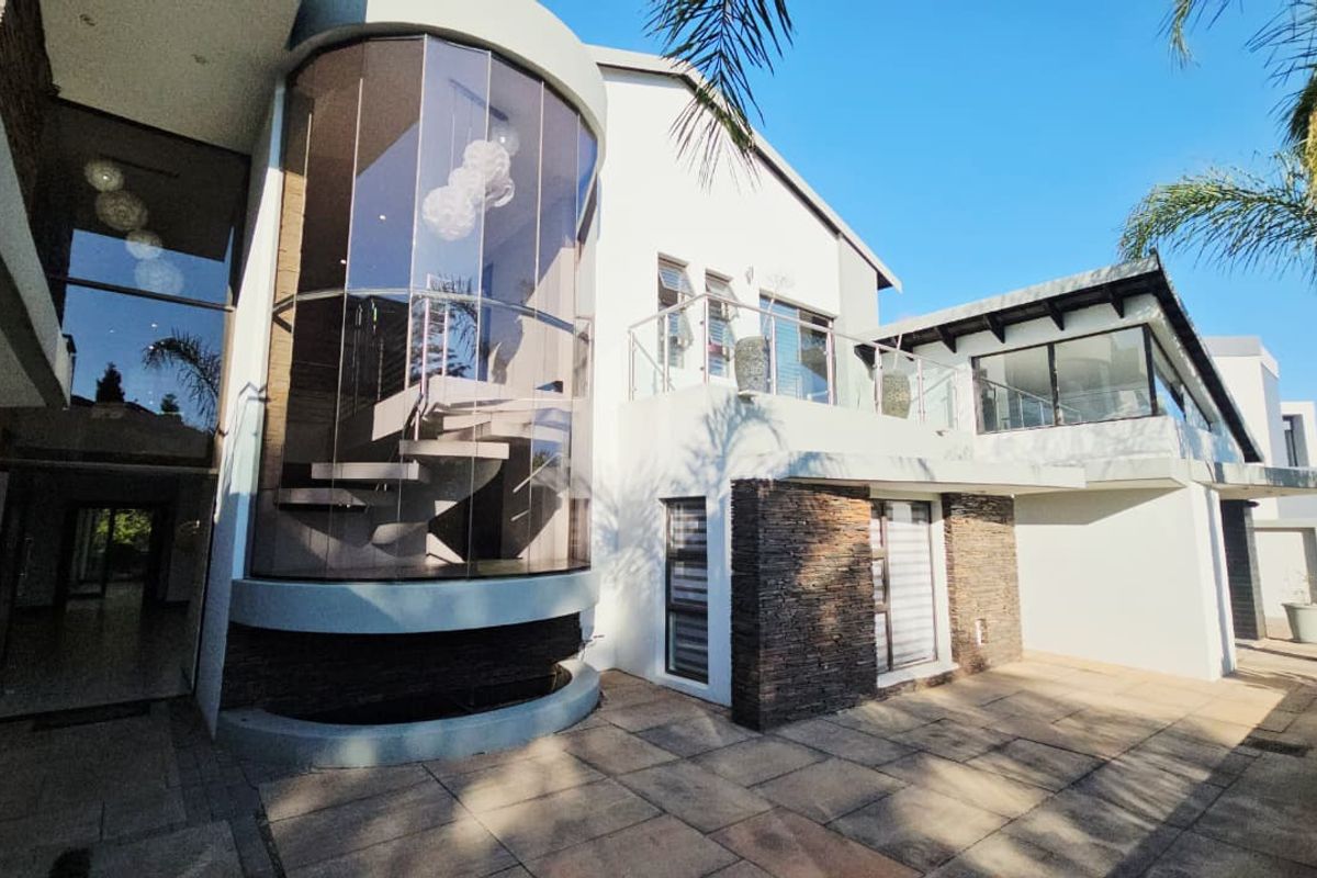 Modern rear exterior featuring a striking curved glass facade, floating staircase, stone cladding, and a spacious paved patio area.