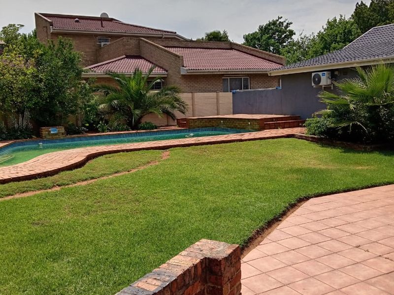 A spacious backyard featuring a swimming pool, tiled patio area, and established garden with brick retaining walls and a multi-level tiled roof house.