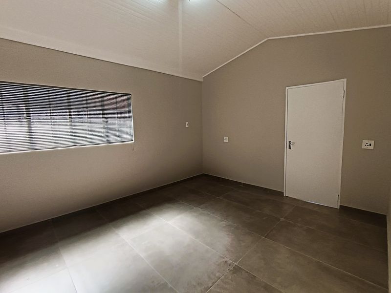 A clean, vacant bedroom featuring neutral wall tones, large format floor tiles, a vaulted ceiling, and fitted venetian blinds.