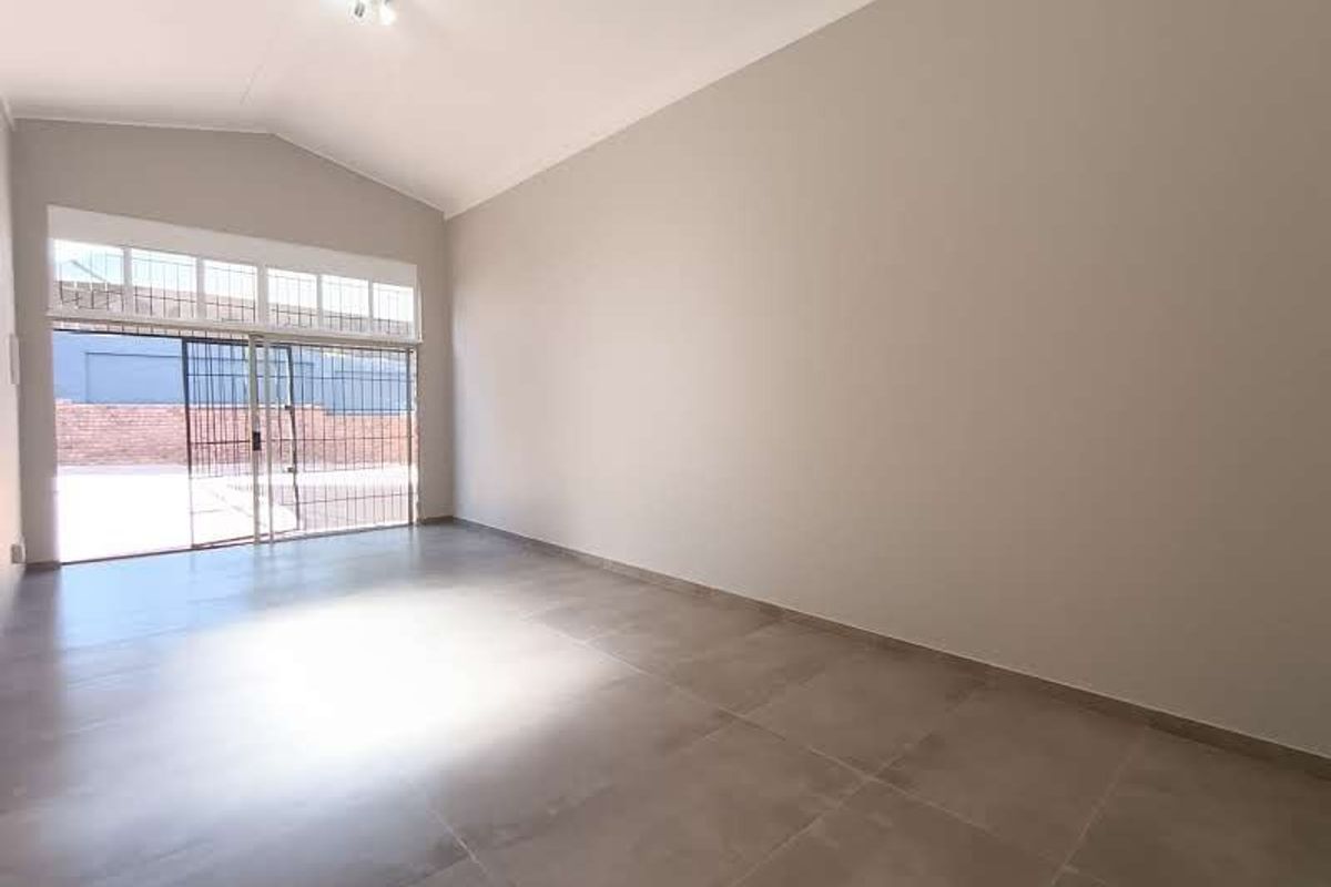 A bright, spacious, and empty commercial living area featuring large-format floor tiles, a vaulted ceiling, and a sliding glass door with security gate.
