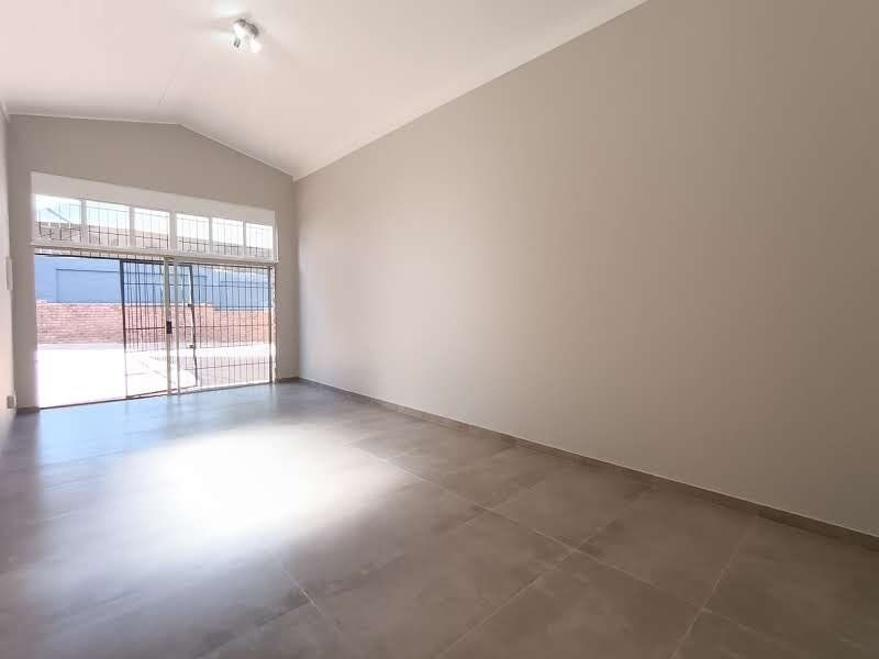 A bright, spacious, and empty commercial living area featuring large-format floor tiles, a vaulted ceiling, and a sliding glass door with security gate.