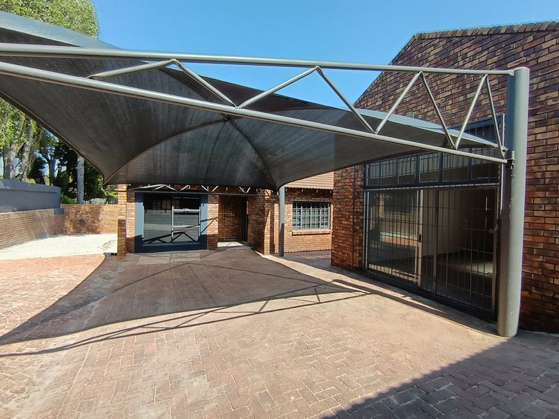 A paved driveway area featuring a large structural shade sail, face brick exterior walls, and secure metal security gates and shutters.