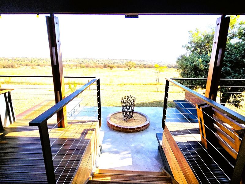 An elevated timber deck featuring a modern steel balustrade and a central brick fire pit, overlooking an expansive natural landscape.