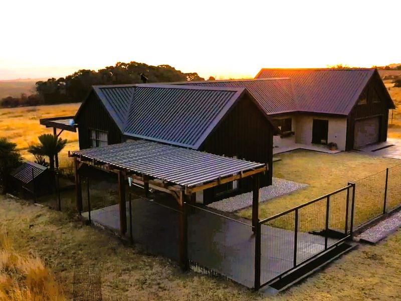 Aerial view of a modern farmhouse-style property featuring a metal roof, timber pergola, and fenced outdoor area set in a rural landscape.