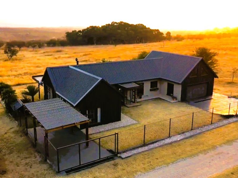 Aerial view of a modern house with a metal roof, covered patio, and perimeter fencing, set in a rural landscape at sunset.