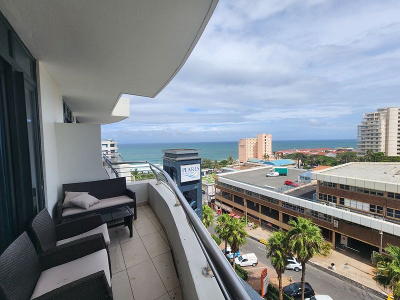 A spacious balcony featuring tiled flooring and a modern stainless steel and glass balustrade, offering expansive coastal and urban views.