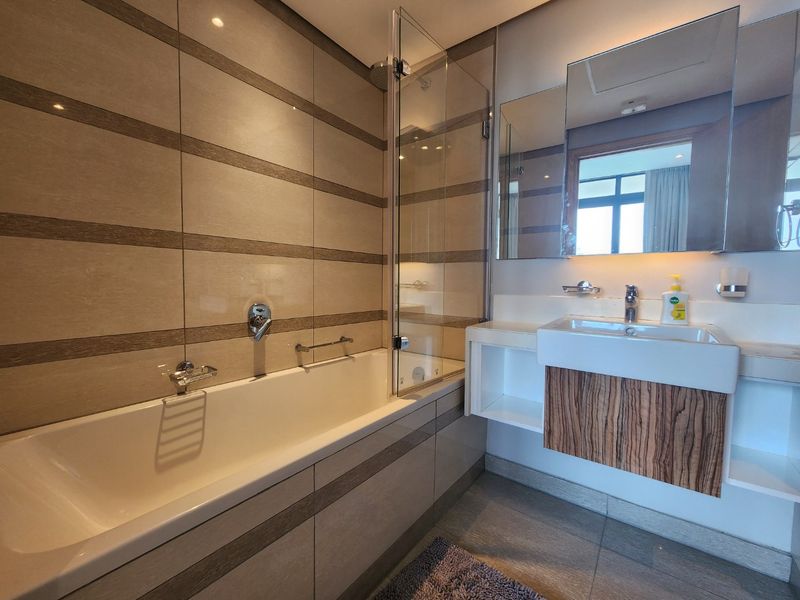Modern bathroom featuring a built-in bathtub with glass screen, wall-mounted vanity with integrated basin, and contemporary tiled finishes.