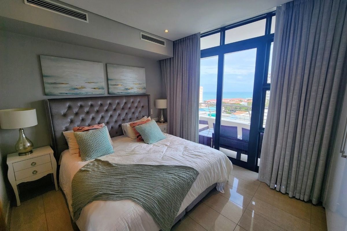 Modern bedroom featuring tiled floors, ducted air conditioning, and large glass sliding doors opening to a balcony with coastal views.