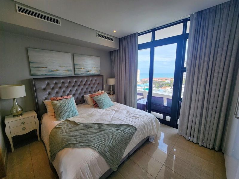 Modern bedroom featuring tiled floors, ducted air conditioning, and large glass sliding doors opening to a balcony with coastal views.