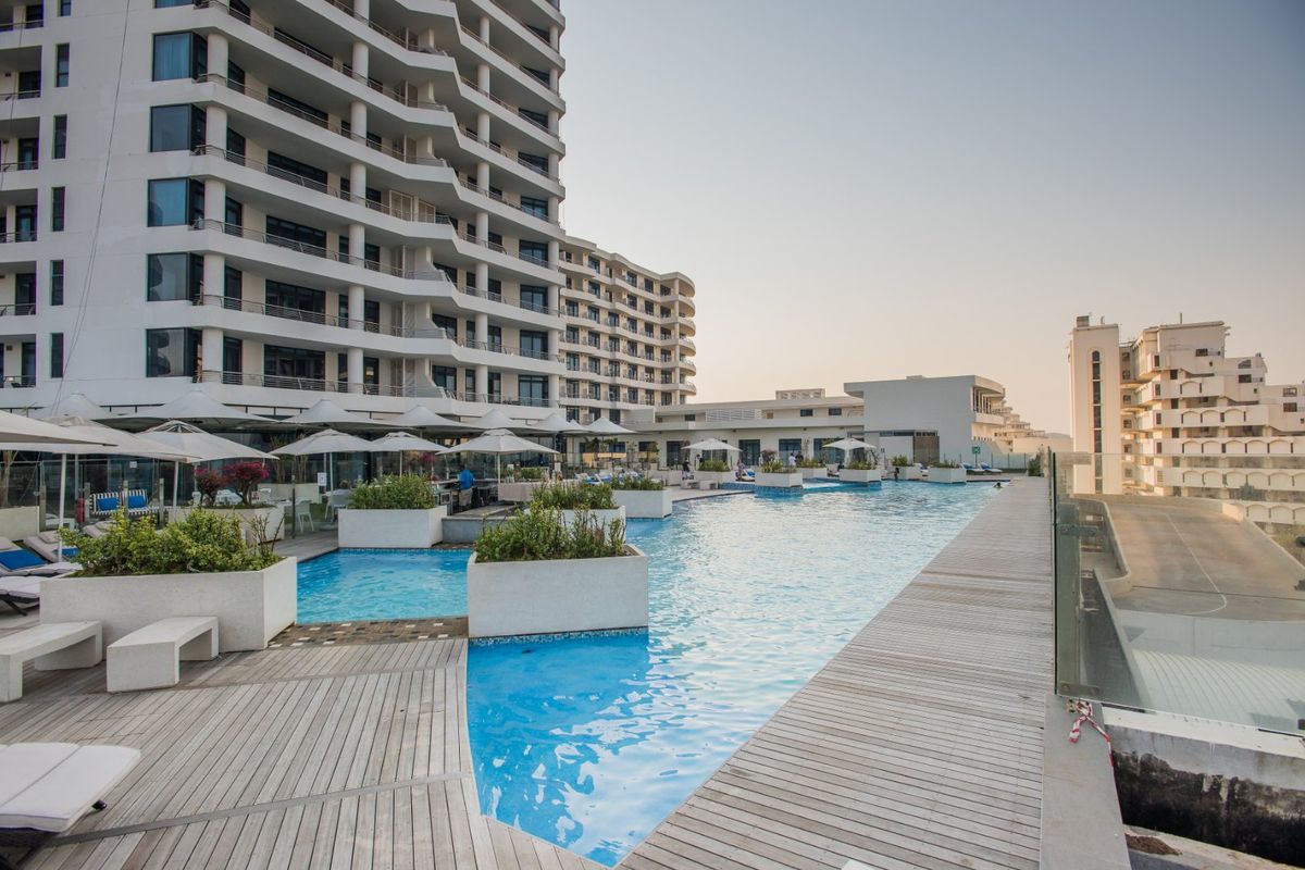 A well-maintained outdoor swimming pool area featuring timber decking, glass balustrades, and integrated concrete planters set against a modern apartment building.