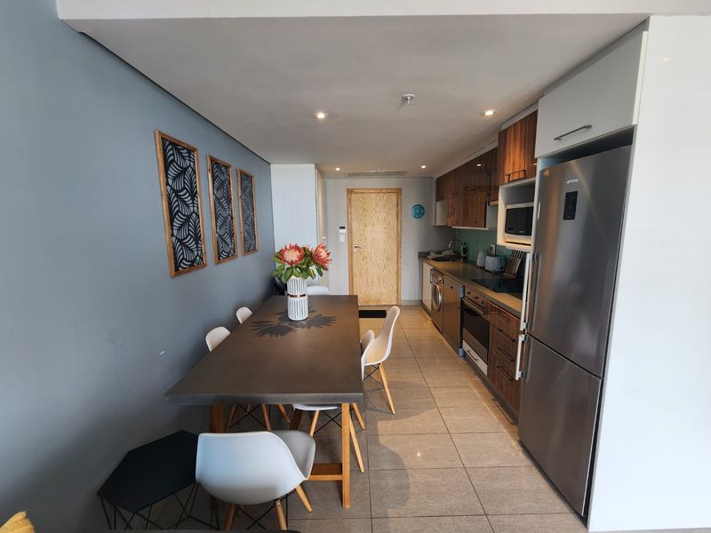 Modern open-plan kitchen and dining area featuring sleek cabinetry, integrated appliances, tiled flooring, and recessed lighting in a contemporary apartment setting.