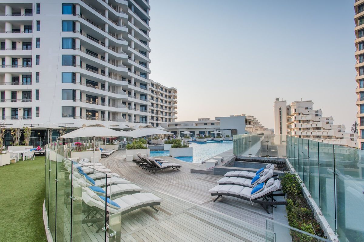 A well-maintained outdoor pool area featuring timber decking, glass balustrades, and artificial turf, set against a modern high-rise apartment complex.