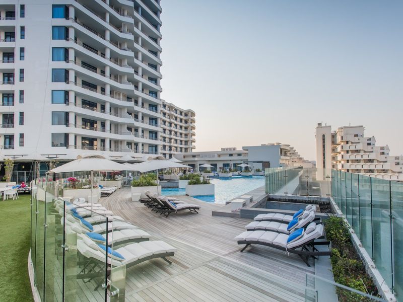 A well-maintained outdoor pool area featuring timber decking, glass balustrades, and artificial turf, set against a modern high-rise apartment complex.