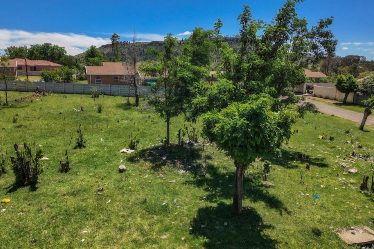 A vacant residential plot featuring a grass lawn, mature trees, and a precast concrete boundary wall in a suburban setting.