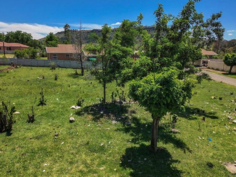 A vacant residential plot featuring a grass lawn, mature trees, and a precast concrete boundary wall in a suburban setting.