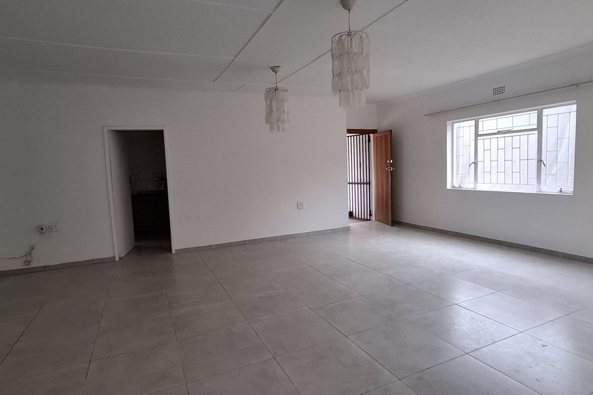 A spacious, empty living area featuring neutral tiled flooring, large windows with security bars, and modern pendant lighting.
