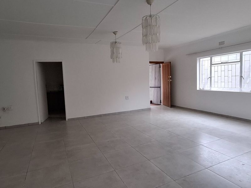 A spacious, empty living area featuring neutral tiled flooring, large windows with security bars, and modern pendant lighting.