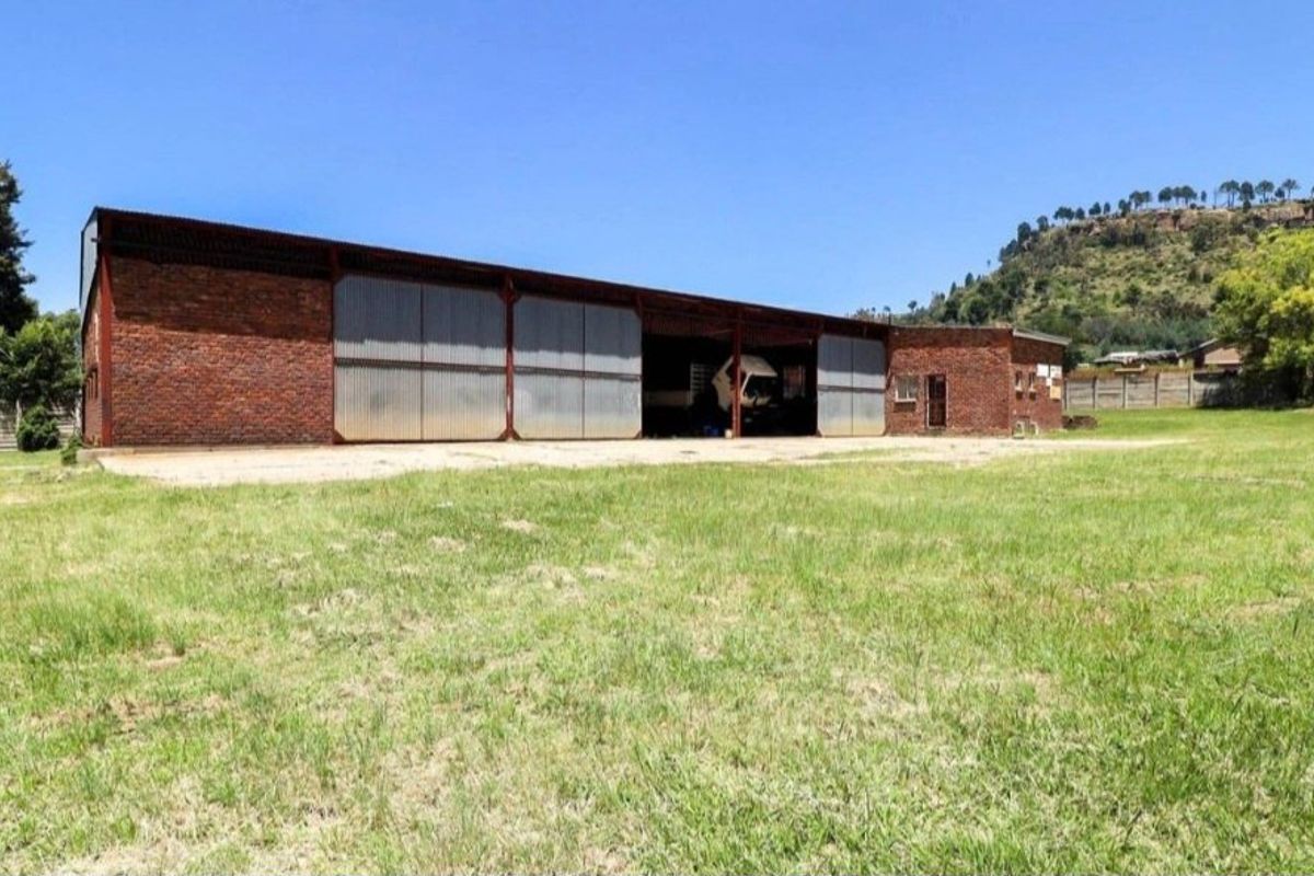 Industrial warehouse building featuring exposed brick walls, corrugated metal sliding doors, and a concrete apron, set on a large open grass plot.
