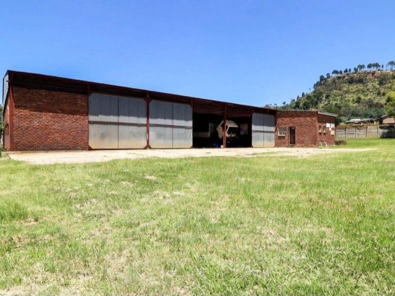 Industrial warehouse building featuring exposed brick walls, corrugated metal sliding doors, and a concrete apron, set on a large open grass plot.
