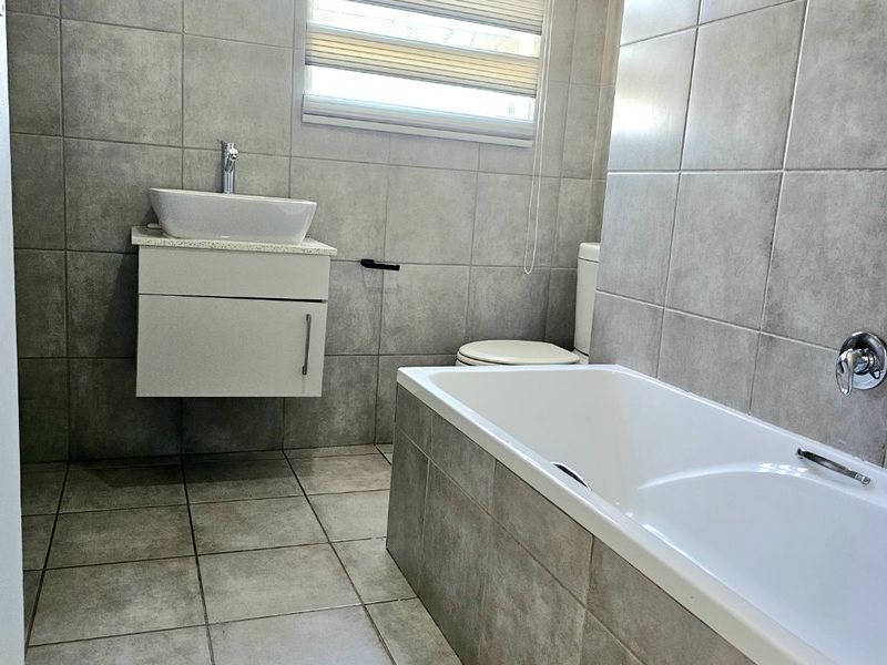 A modern, well-maintained bathroom featuring grey ceramic tiling, a wall-mounted vanity with a vessel sink, and a built-in bathtub.