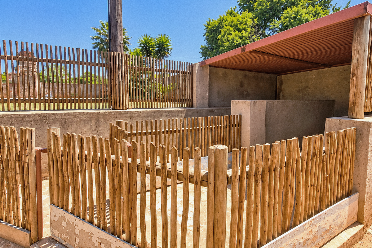 An outdoor utility enclosure featuring wooden pole fencing, concrete retaining walls, and a covered section with a corrugated metal roof.