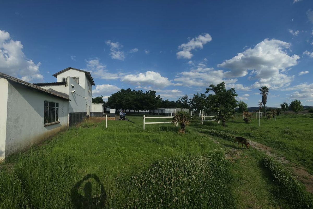 A wide-angle view of the farm property exterior, featuring white masonry buildings, post and rail fencing, and expansive green grounds under a clear sky.