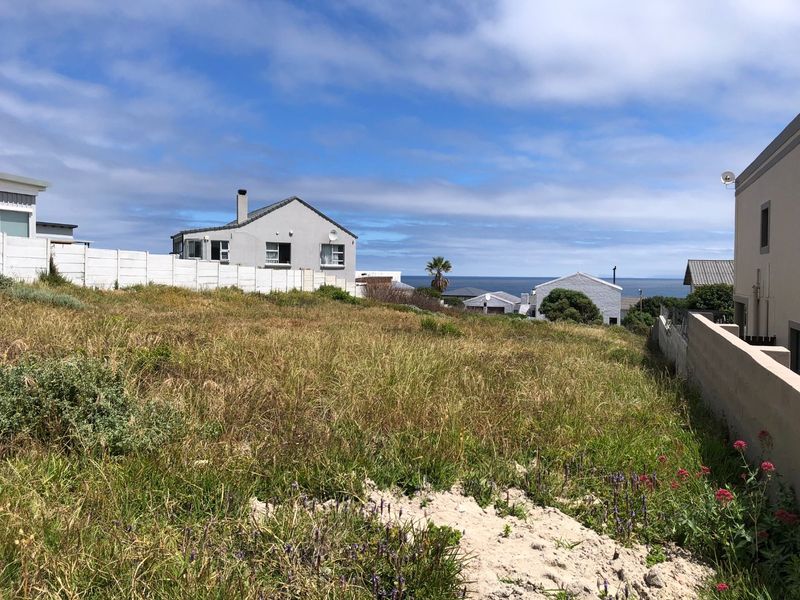 A vacant residential plot featuring natural coastal vegetation, sandy terrain, and a precast concrete boundary wall with distant ocean views.