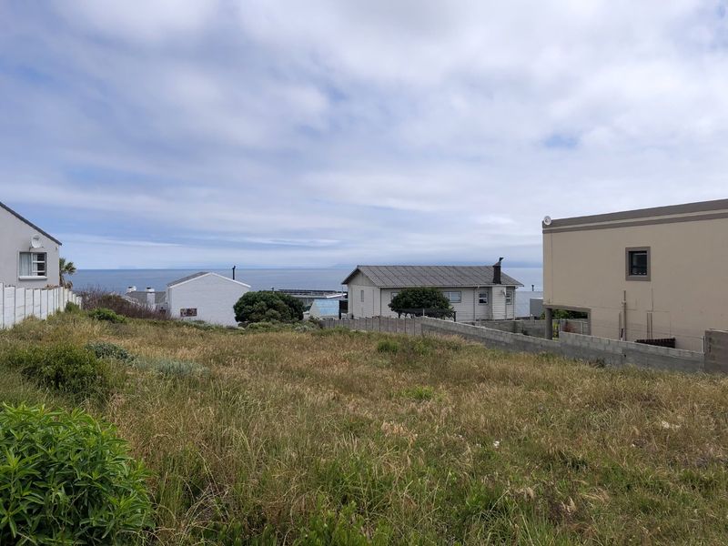 A vacant residential plot featuring natural grass cover, enclosed by boundary walls and fencing, with surrounding neighborhood homes and distant ocean views.