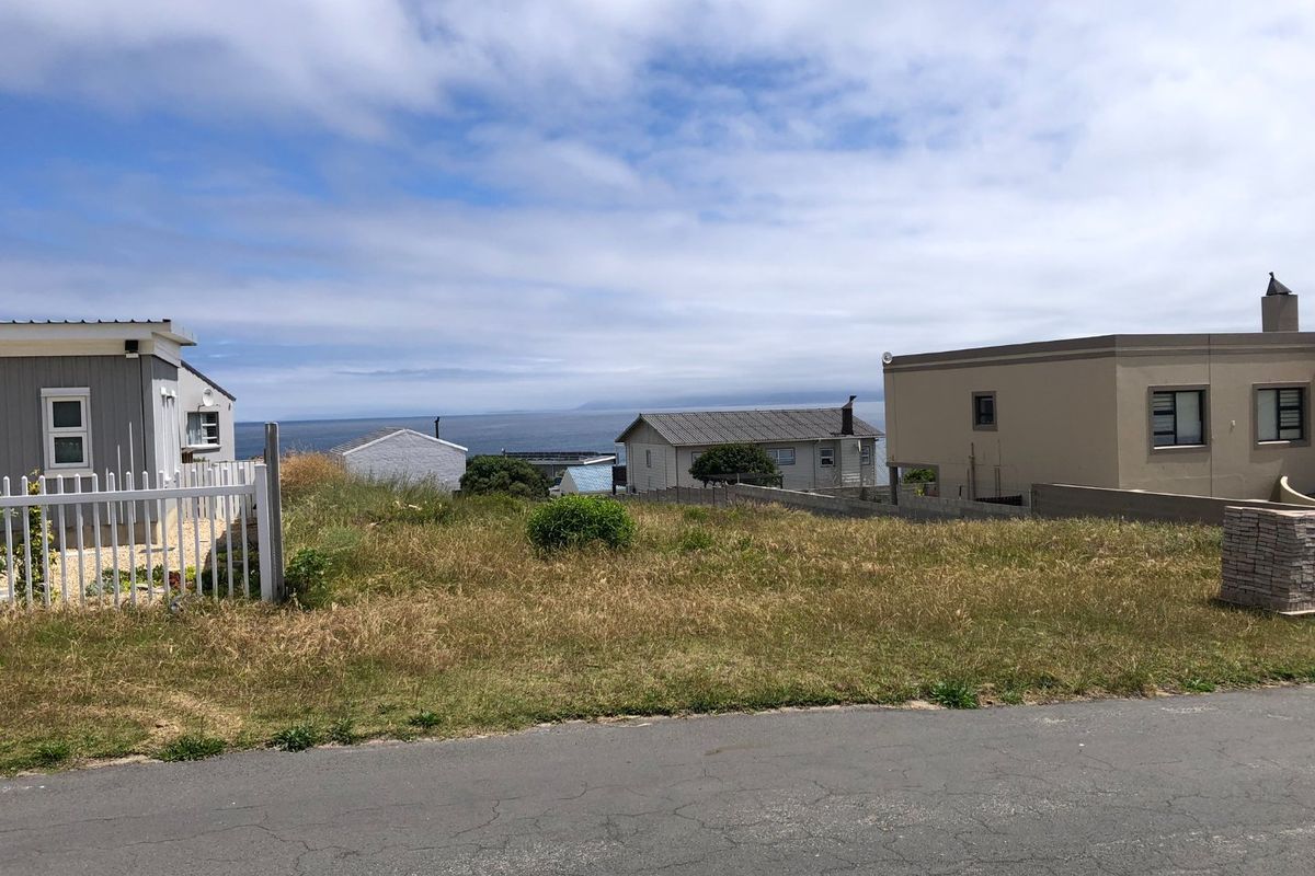 A vacant residential plot situated along a paved road, offering a clear view of the surrounding neighborhood and distant ocean horizon.