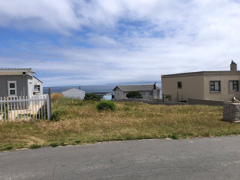 A vacant residential plot situated along a paved road, offering a clear view of the surrounding neighborhood and distant ocean horizon.