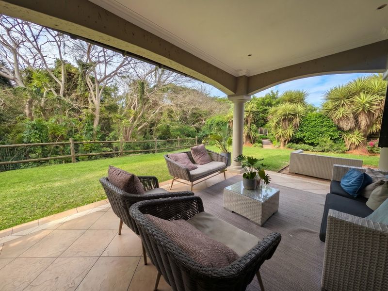 A spacious covered patio featuring tiled flooring and support columns, overlooking a lush private garden and natural greenery.