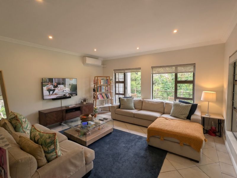 A bright and well-maintained living room featuring tiled flooring, recessed downlighting, split-system air conditioning, and large windows with views of the surrounding greenery.