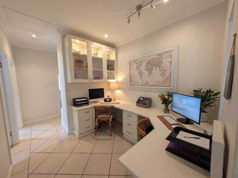 A well-appointed home office featuring custom built-in desk cabinetry, glass-fronted display storage, tiled flooring, and modern track lighting.