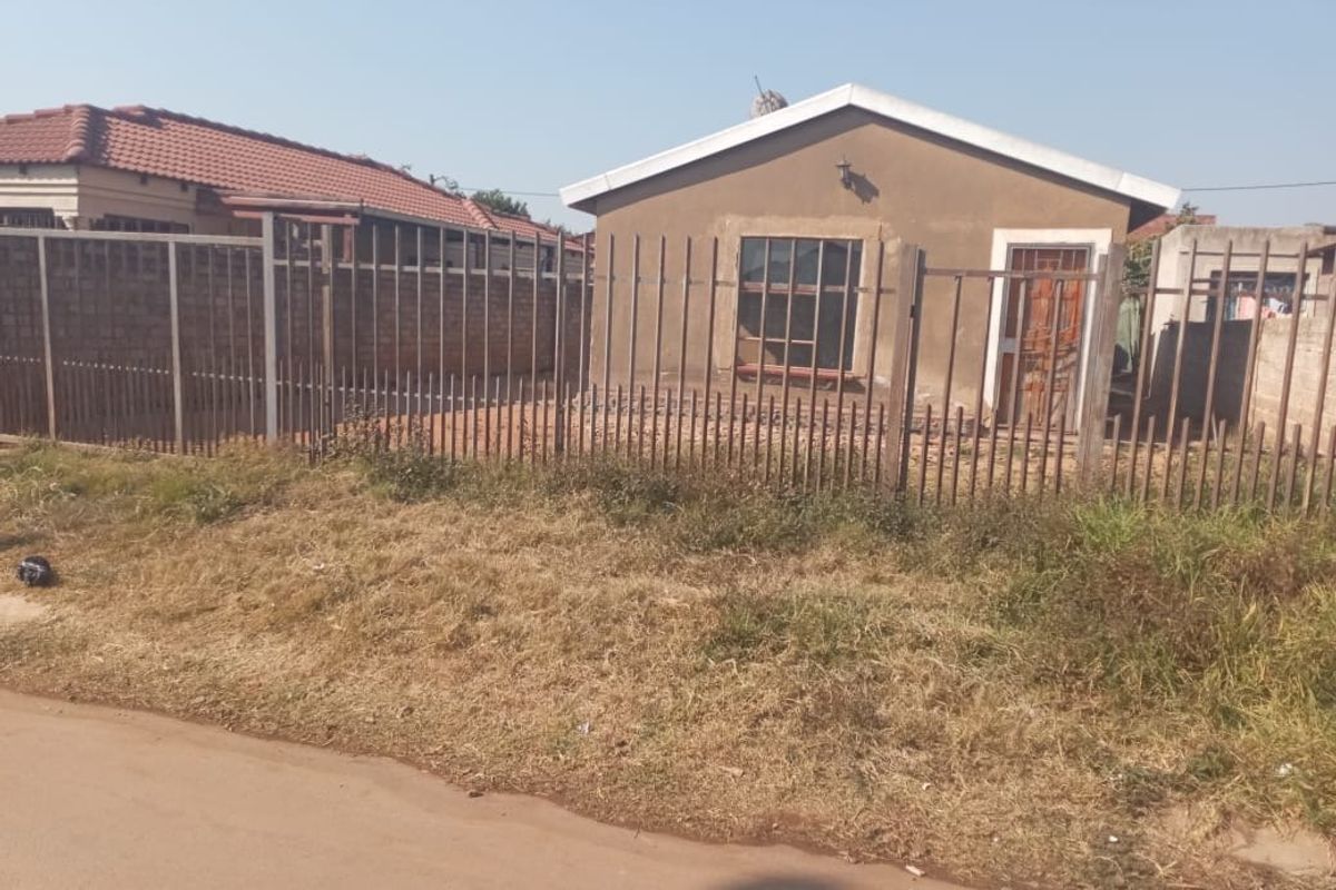 Front view of a single-story residential house featuring a tiled roof, plastered exterior walls, and secure metal perimeter fencing.