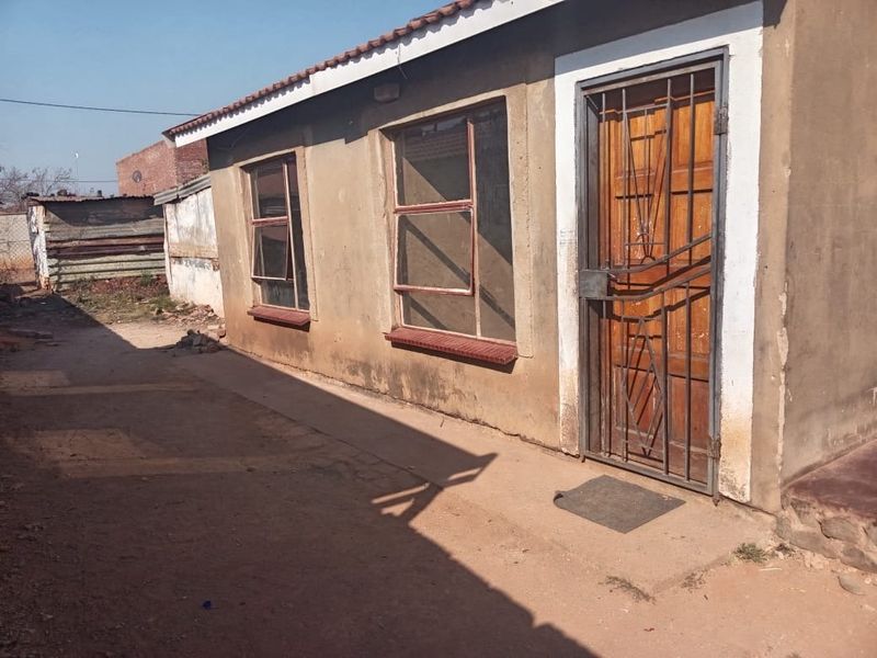 Exterior view of a residential property featuring a metal security gate, steel frame windows, and a tiled roof with visible wear to the exterior plaster.
