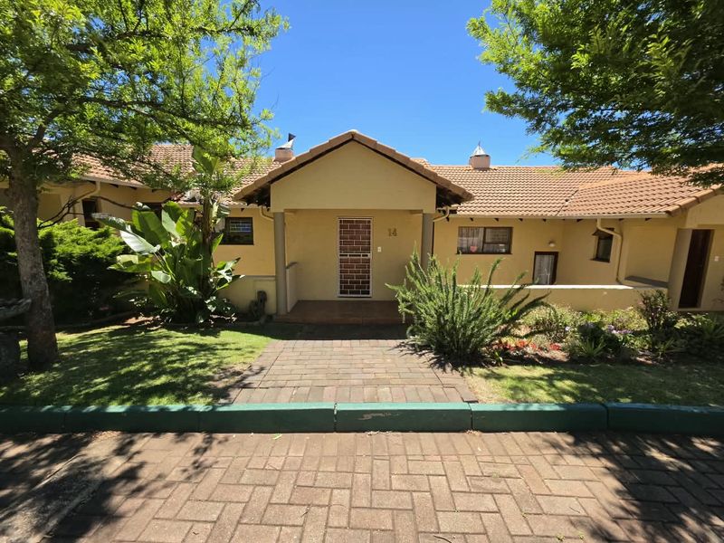 Front elevation of a townhouse featuring a tiled roof, covered entrance porch, security gate, and a well-maintained garden with a brick paved driveway.