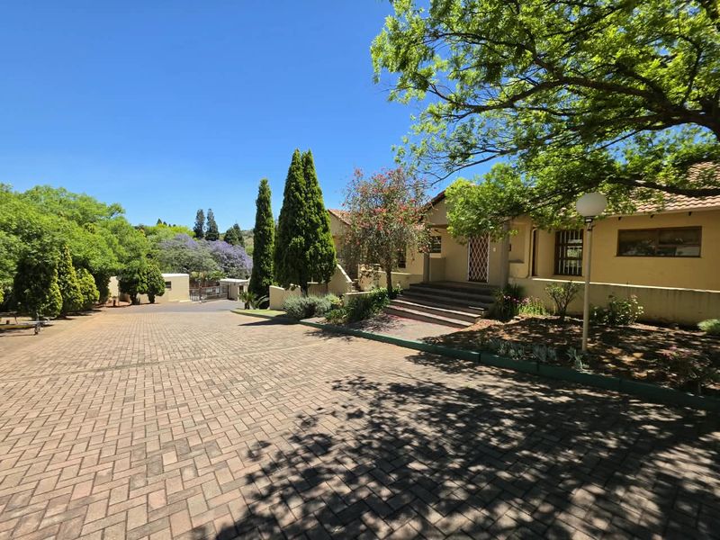 A well-maintained front exterior of a townhouse featuring a spacious paved driveway, mature garden landscaping, and a secure entrance with steps.