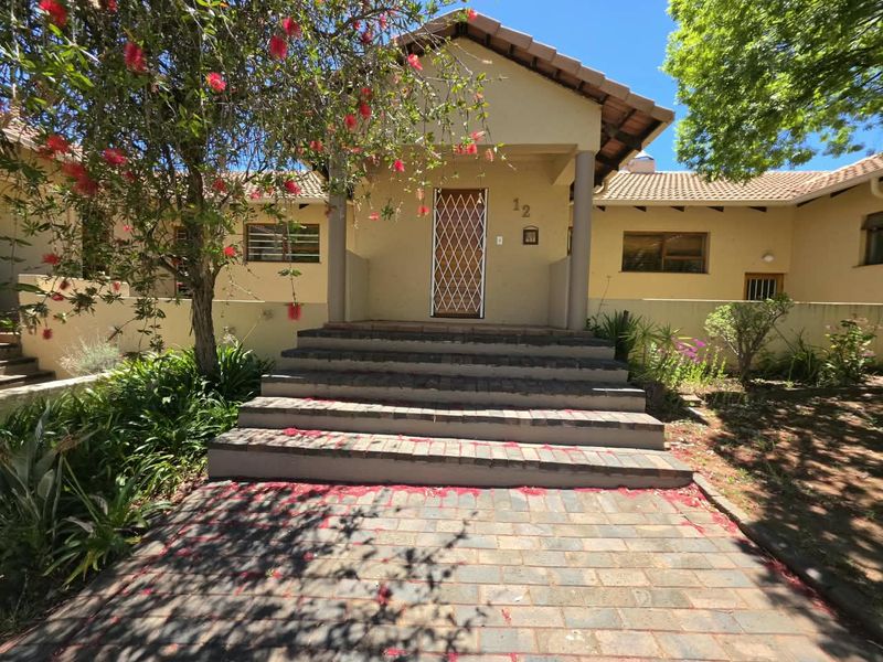 Front entrance of a townhouse featuring a paved walkway, brick stairs, and a security gate, set within a landscaped garden area.