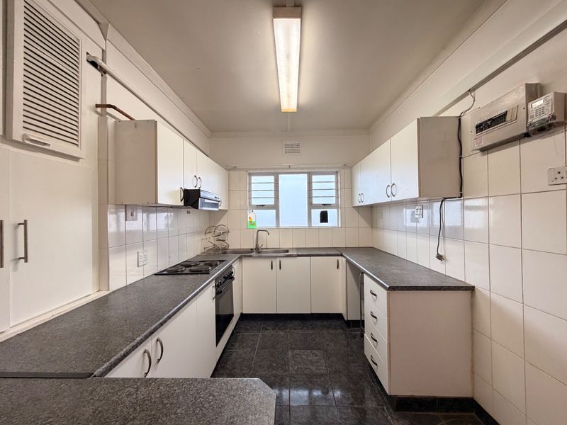 A functional, galley-style kitchen featuring white cabinetry, dark laminate countertops, tiled splashbacks, and built-in cooking appliances, offering a clean and practical workspace.