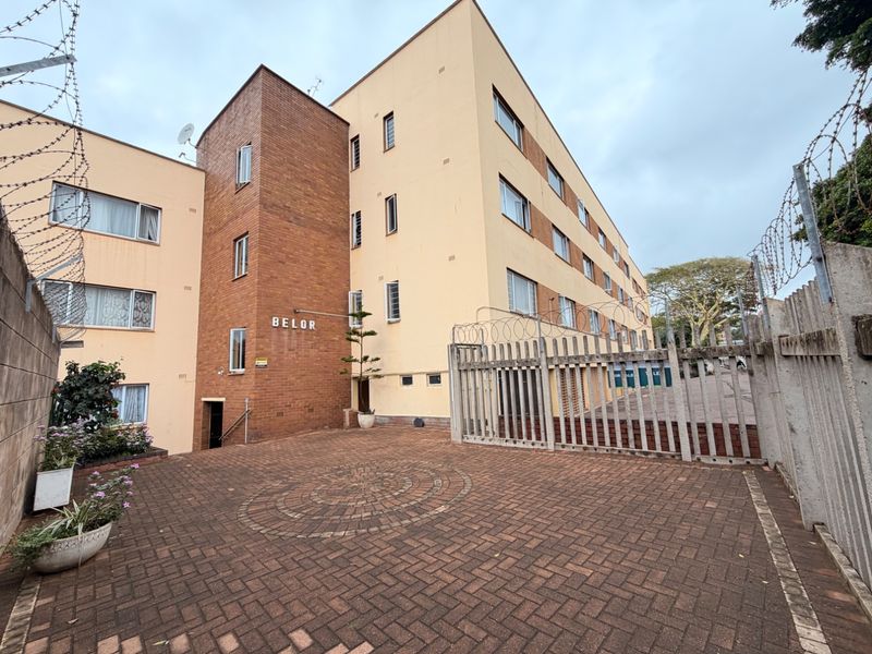 Exterior view of the Belor apartment building featuring a paved entrance courtyard, secure metal gate, and perimeter security fencing.