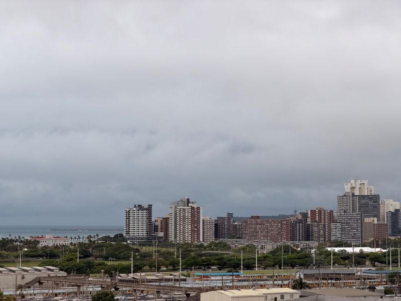 An elevated cityscape view overlooking urban buildings, infrastructure, and coastal surroundings under a cloudy sky.