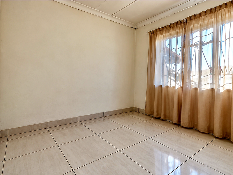 A bright, empty bedroom featuring neutral tiled flooring, clean walls, and a window fitted with security bars.