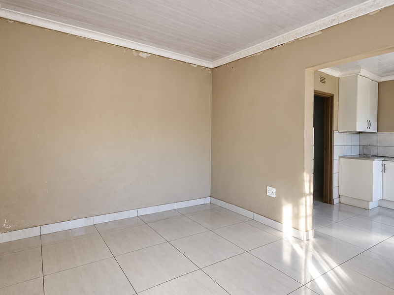 An empty open-plan living area featuring tiled floors and a view into a compact kitchen with built-in cabinetry and tiled backsplash.