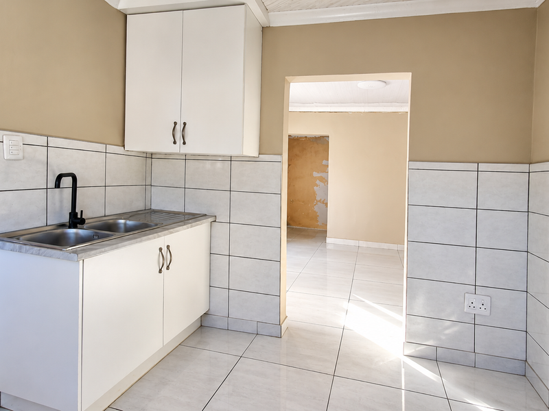A compact kitchen area featuring white cabinetry, a stainless steel sink with a modern black tap, and tiled finishes, leading into an adjacent living space.