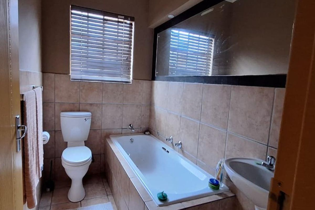 A functional bathroom featuring a built-in bathtub, pedestal basin, toilet, and wall-to-wall mirror with neutral tiled finishes.