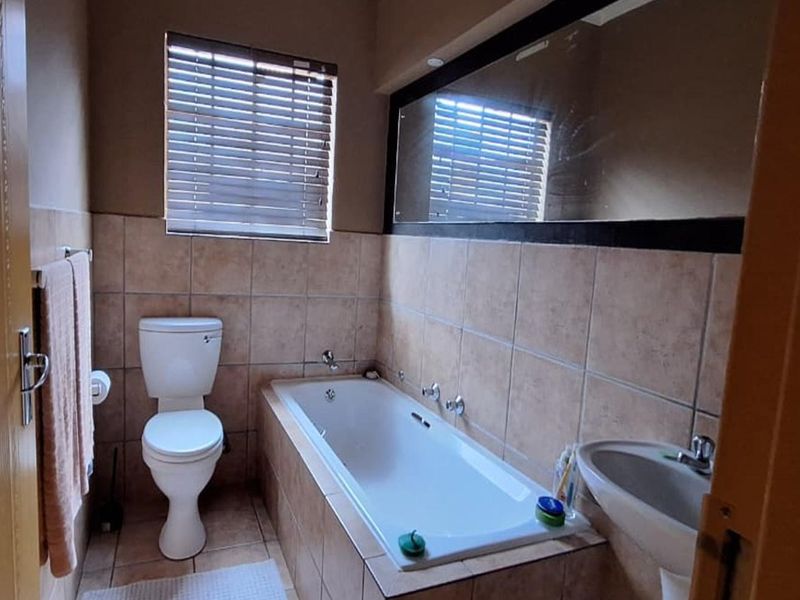 A functional bathroom featuring a built-in bathtub, pedestal basin, toilet, and wall-to-wall mirror with neutral tiled finishes.