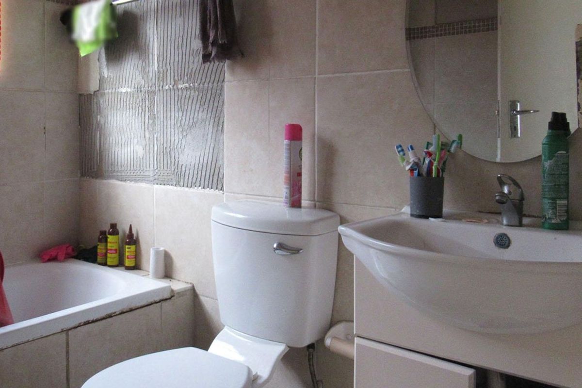 A bathroom featuring a bathtub, toilet, and vanity basin, currently requiring repair due to missing wall tiles and exposed adhesive.